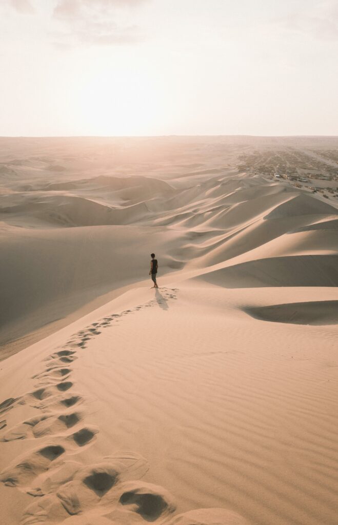 A lone traveler walks across the vast sand dunes of Ica, Peru, under the warm glow of a sunset.